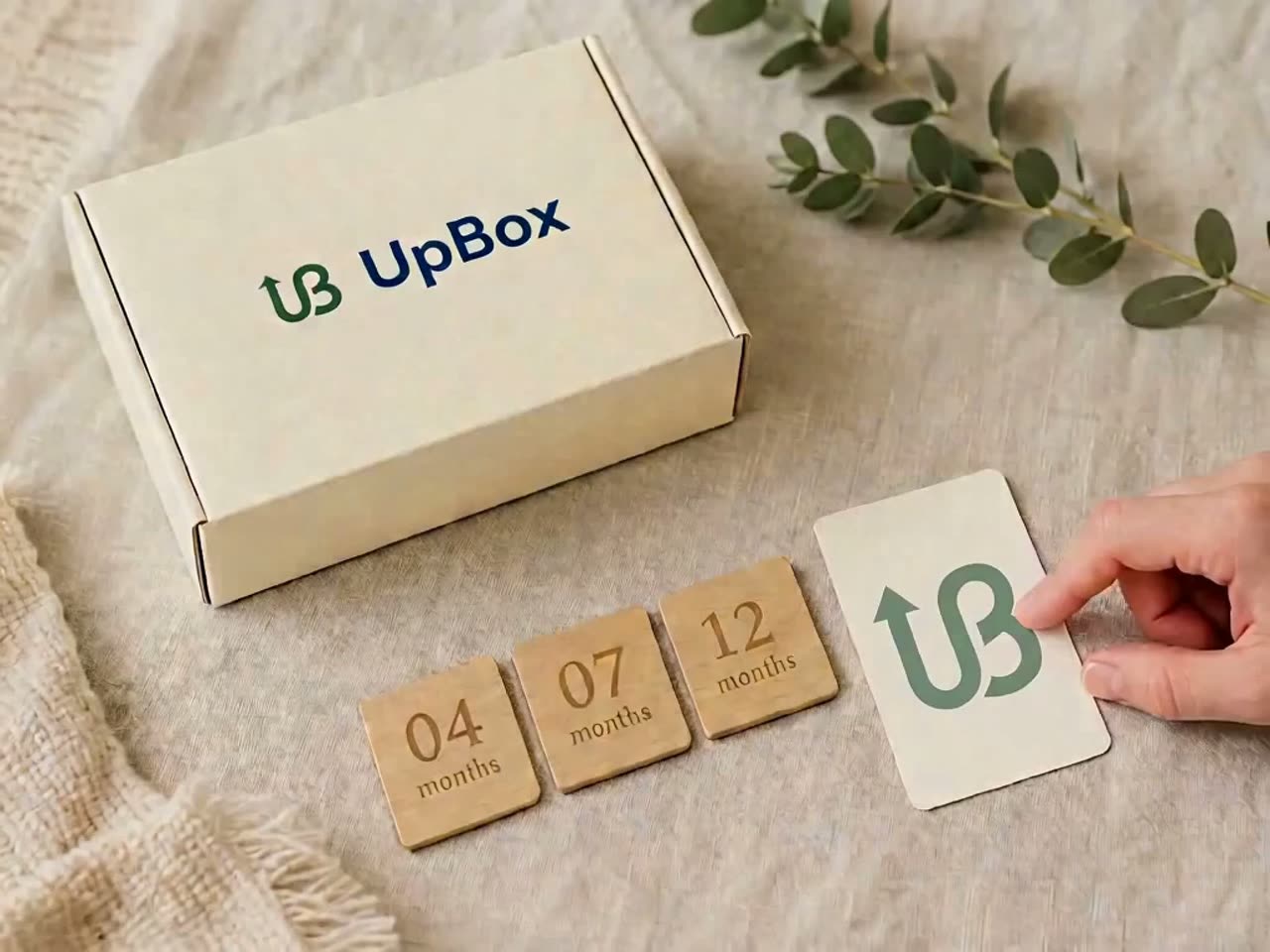 A hand tapping wooden age tiles beside a closed cream UpBox box printed with the green UB monogram and navy UpBox wordmark